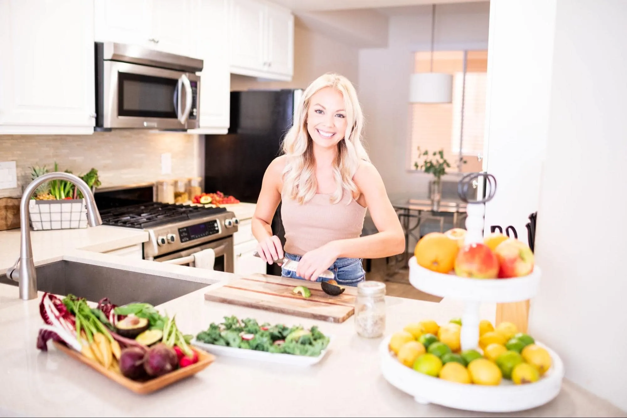 A woman with long blonde hair is standing in a modern kitchen with white cabinets, cutting vegetables on a wooden board. The countertop has various fruits and greens, and there are shelves of fresh produce in the foreground. She is smiling at the camera.