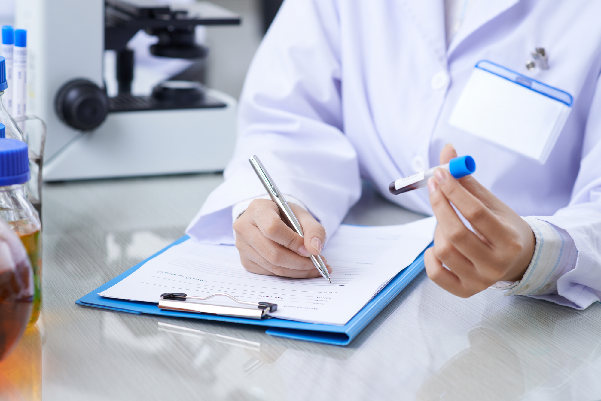A person in a lab coat holding a test tube with a blue cap in their left hand and writing on a clipboard with their right. A microscope and various laboratory equipment are visible in the background, meticulously documenting dna exoneration statistics for an important research project.